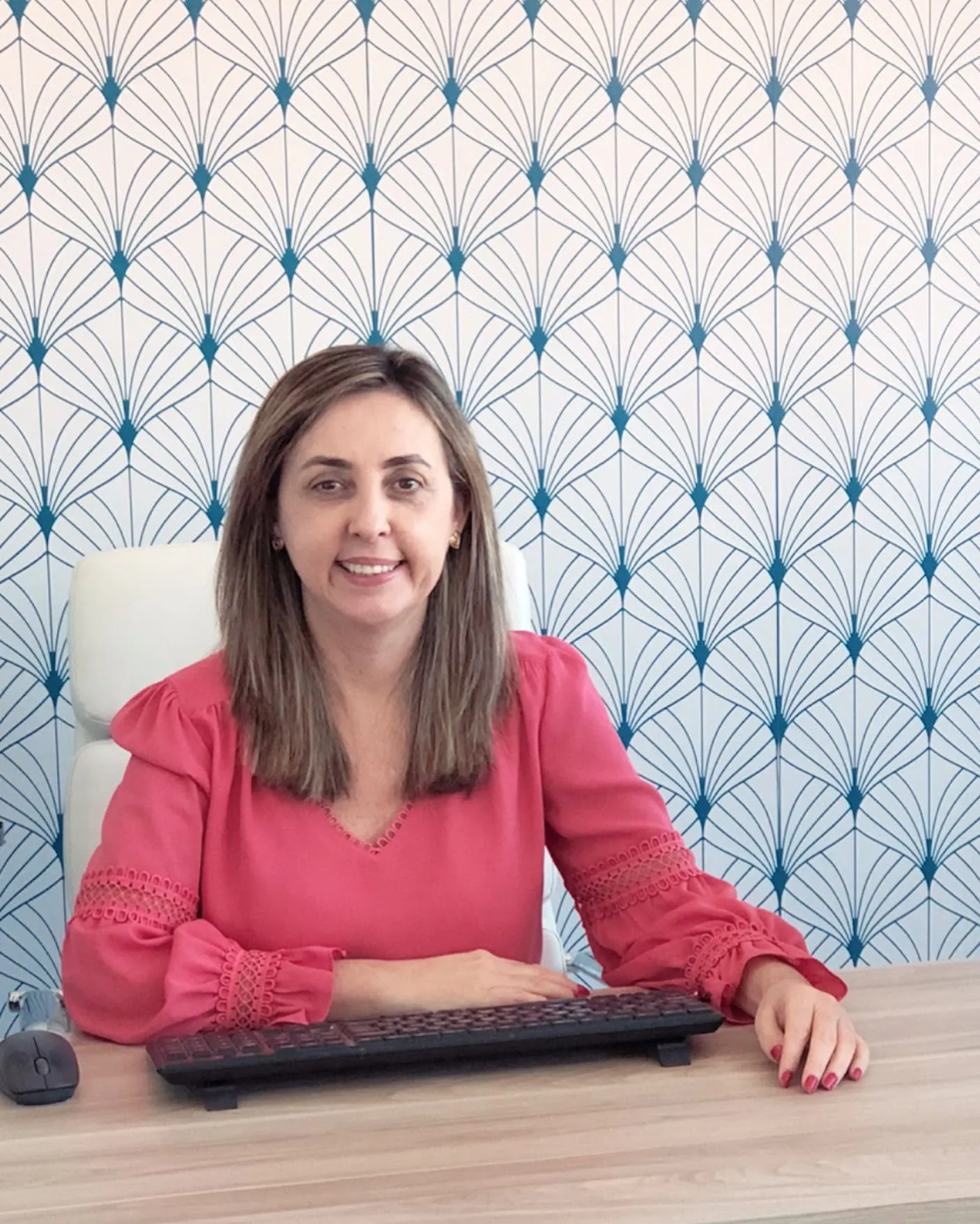 Vânia Beletate sorrindo à mesa do consultório, com o papel de parede de leques ao fundo